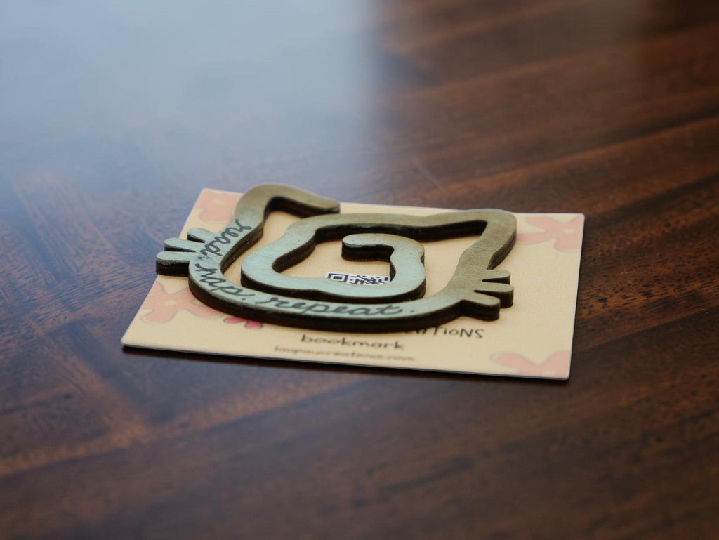 La Apau Creations laser-cut wooden kitty bookmark with read nap repeat text on a branded display card